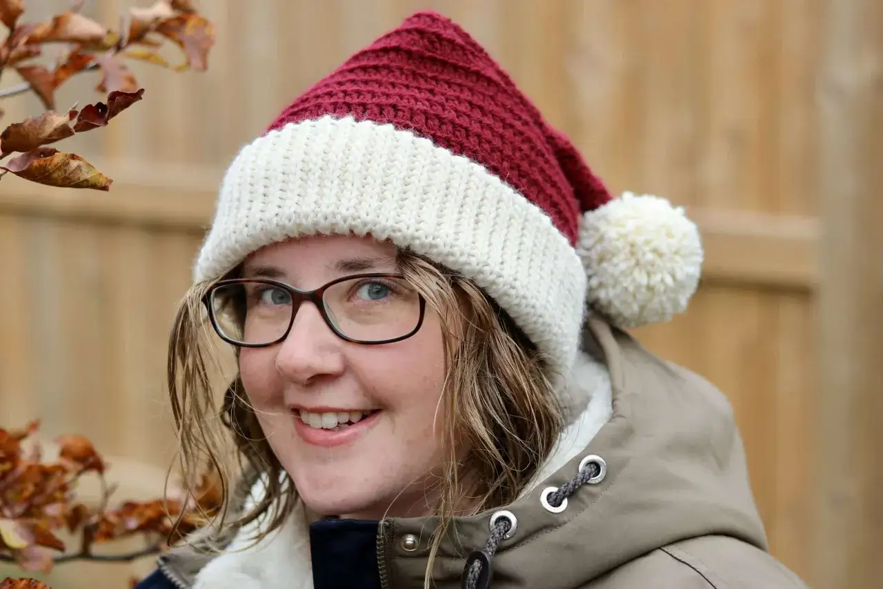 En glad person med glas&ouml;gon b&auml;r en r&ouml;d och vit virkad tomteluva med en fluffig pompom.