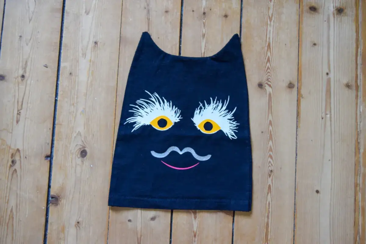 En svart tröja med ett roligt kattansikte, som om det vore en sy sommarskuggan. Ögonen är stora och gula med vita fransar, och en grå mustasch och rosa mun fulländar uttrycket.