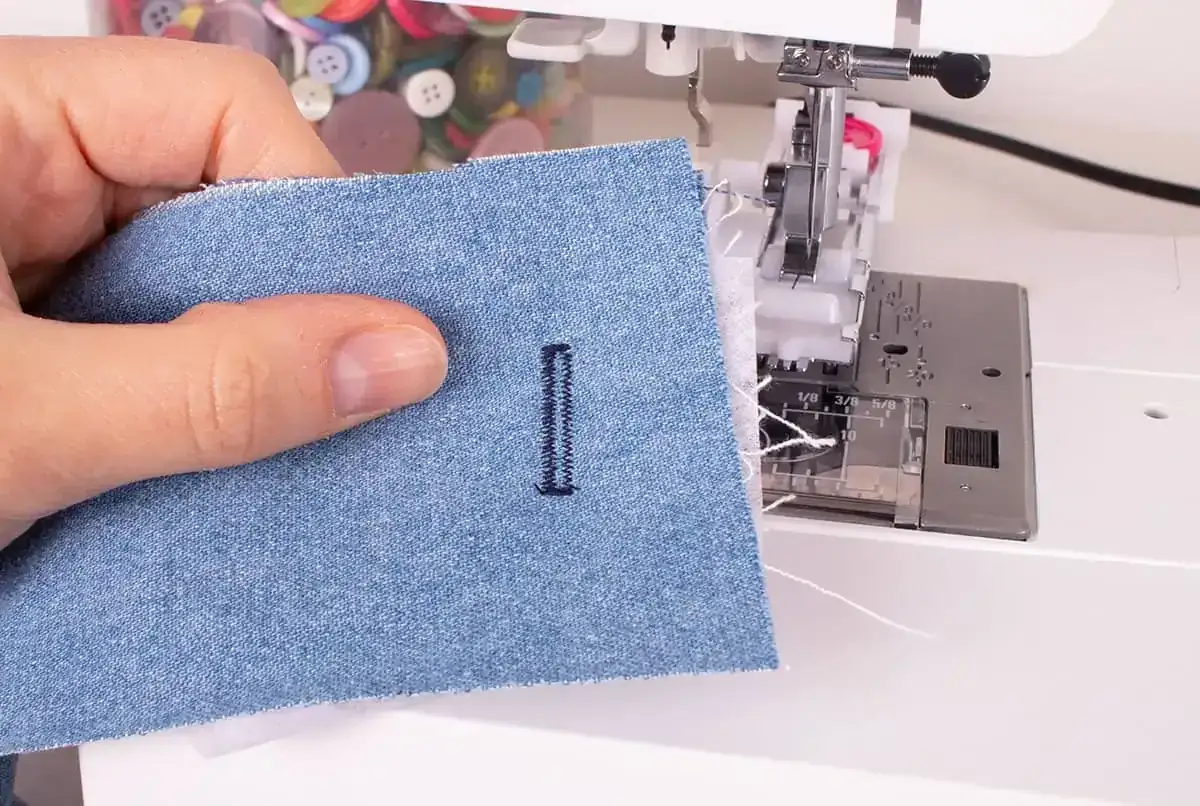 En hand håller upp ett jeansstycke där en symaskin precis har sytt ett knapphål.