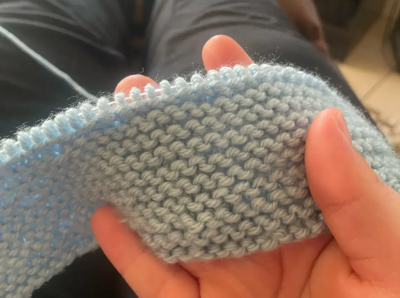 H&auml;nder som h&aring;ller i ett ljusbl&aring;tt stickat tyg. M&ouml;nstret &auml;r stockinette stitch svenska, med tydliga rader av r&auml;ta och aviga maskor.