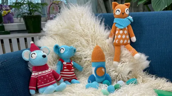 En virkad amigurumi katt med orange och vita prickar sitter på en lurvig matta bredvid en blå mus med tomteluva och en blå elefant i hängselbyxor.