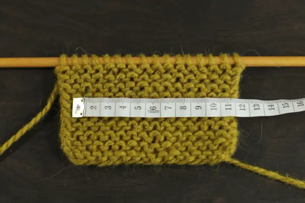 Knitted swatch with a measuring tape across it, showing stitches on wooden needles.