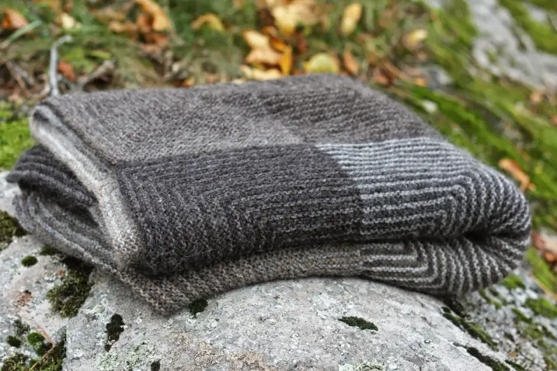 En hopvikt, handstickad filt i gråa nyanser ligger på en mossig sten. Perfekt för mysiga höstkvällar.