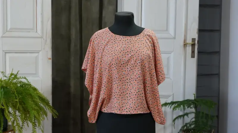 En persikofärgad blus med ett litet mönster av mörka prickar och små blommor. Blusen har en vid passform och korta, fladdermusliknande ärmar.